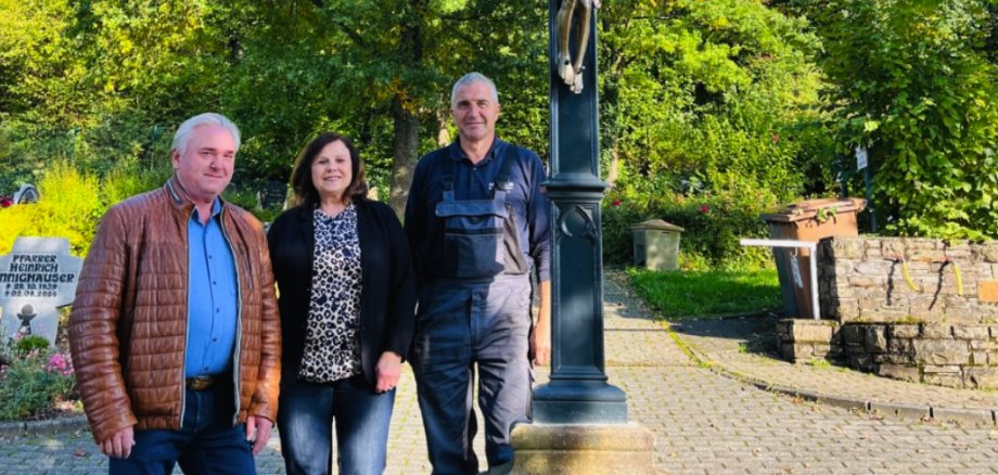 drei Menschen stehen vor einem restaurierten Friedhofskreuz